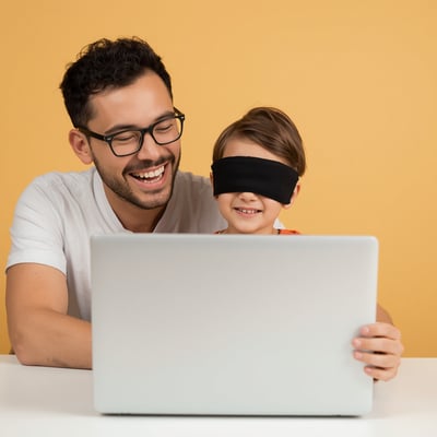 A happy parent and child around 6-8 years old at a plain silver laptop with blank lid and no logos or brand marks, with the child wearing a black blindfold mask, both smiling and having fun together